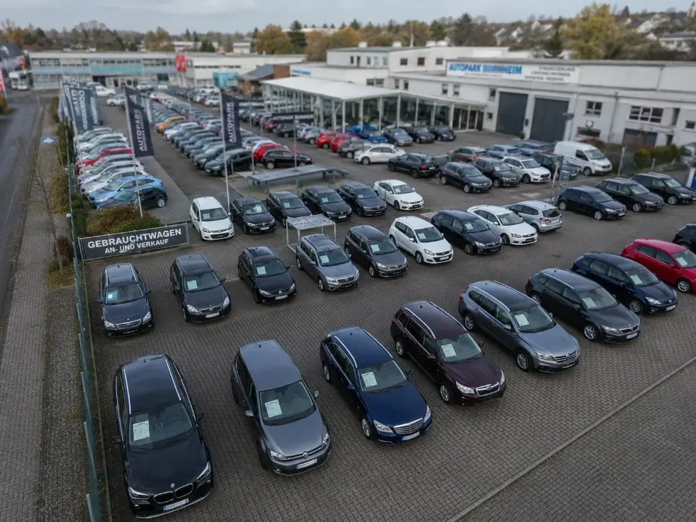 Drohnen-Übersichtsaufnahme eines Gebrauchtwagen-Handels. Aus der Vogelperspektive sind etwa 80 geordnet aufgestellte Fahrzeuge sichtbar, das Verkaufsgebäude mit großer Glasfront und Fahnenmasten von 'Autopark Bornheim'. Im Hintergrund weitere Gewerbegebäude und Vegetation.