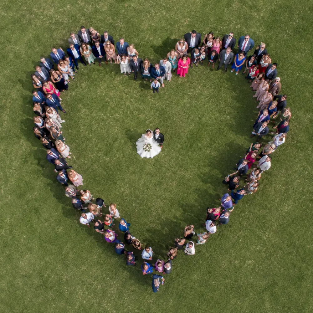 Drohnen-Aufnahme aus der Vogelperspektive: Hochzeitspaar steht im Zentrum einer Wiese, die Braut in weißem Brautkleid mit Brautstrauß, der Bräutigam im dunklen Anzug. Um das Paar bilden alle Hochzeitsgäste eine perfekte Herzform – mehrere Dutzend festlich gekleidete Personen, die nach oben in die Kamera blicken.